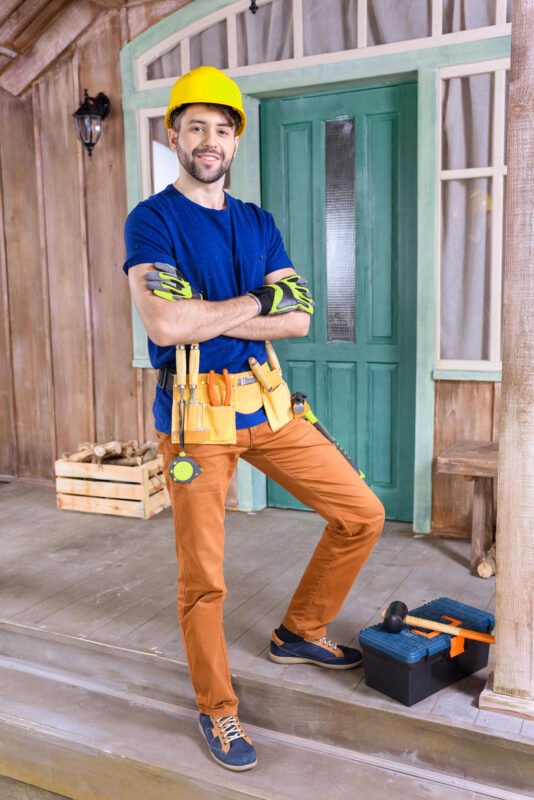 Smiling carpenter in helmet with building tools standing on porch and looking to camera